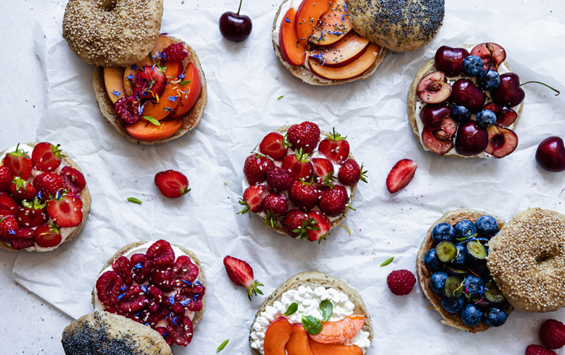Plusieurs bagels sans gluten de la boulangerie Piaf Gluten Free garnis de fruits rouges, fraises, framboises, cerises ainsi que de pêches et d'abricots tranchés